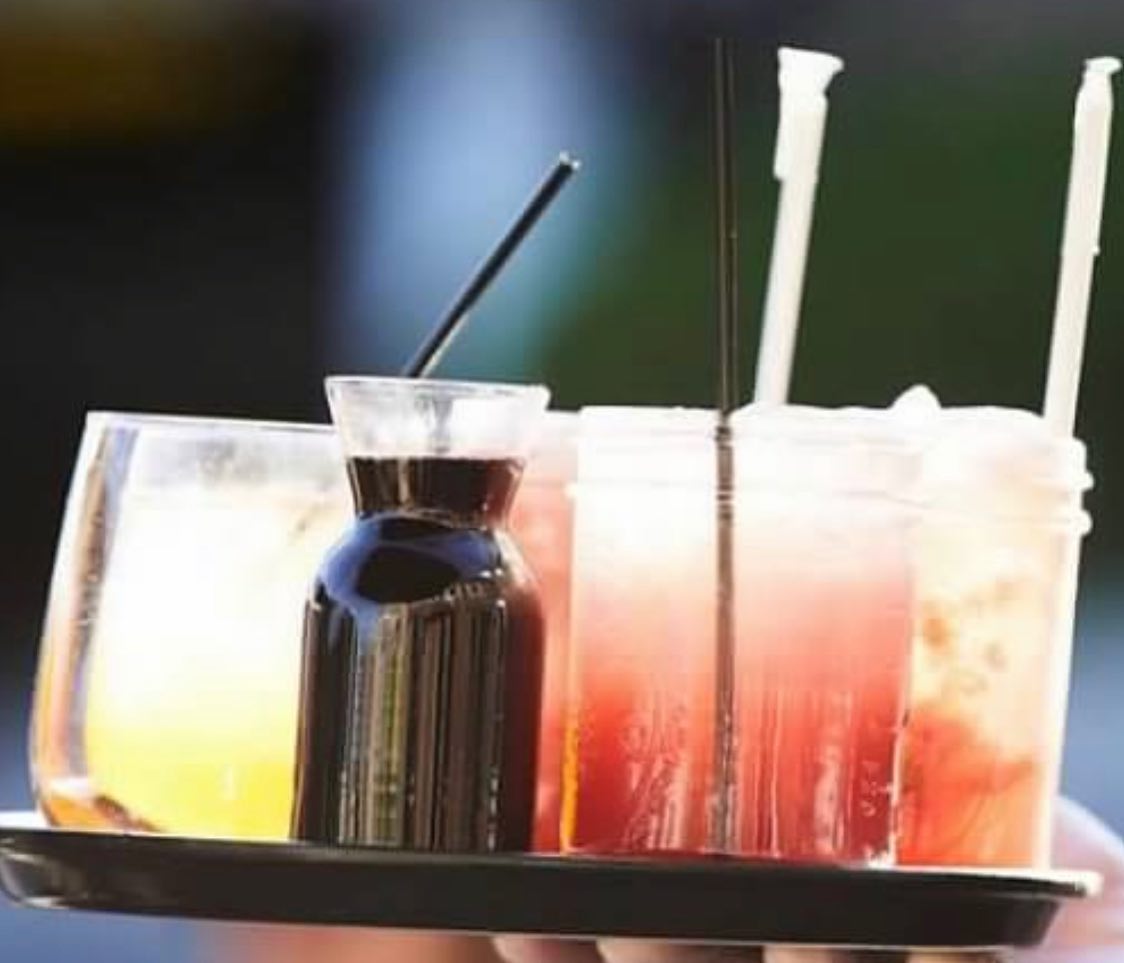 A tray holding a glass of yellow liquid, a dark bottle, and several glasses of pink drinks with straws.