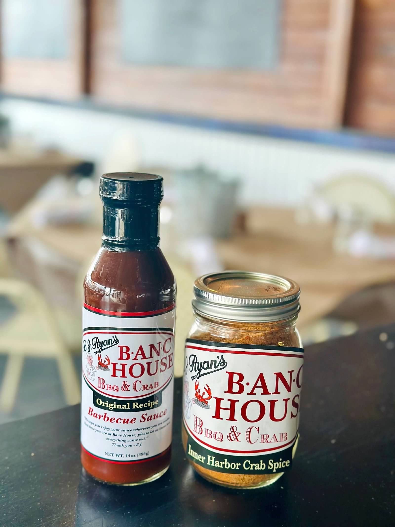 Two Ranch House brand containers, a red barbecue sauce bottle and a seasoning jar, sit on a table in a blurred restaurant.