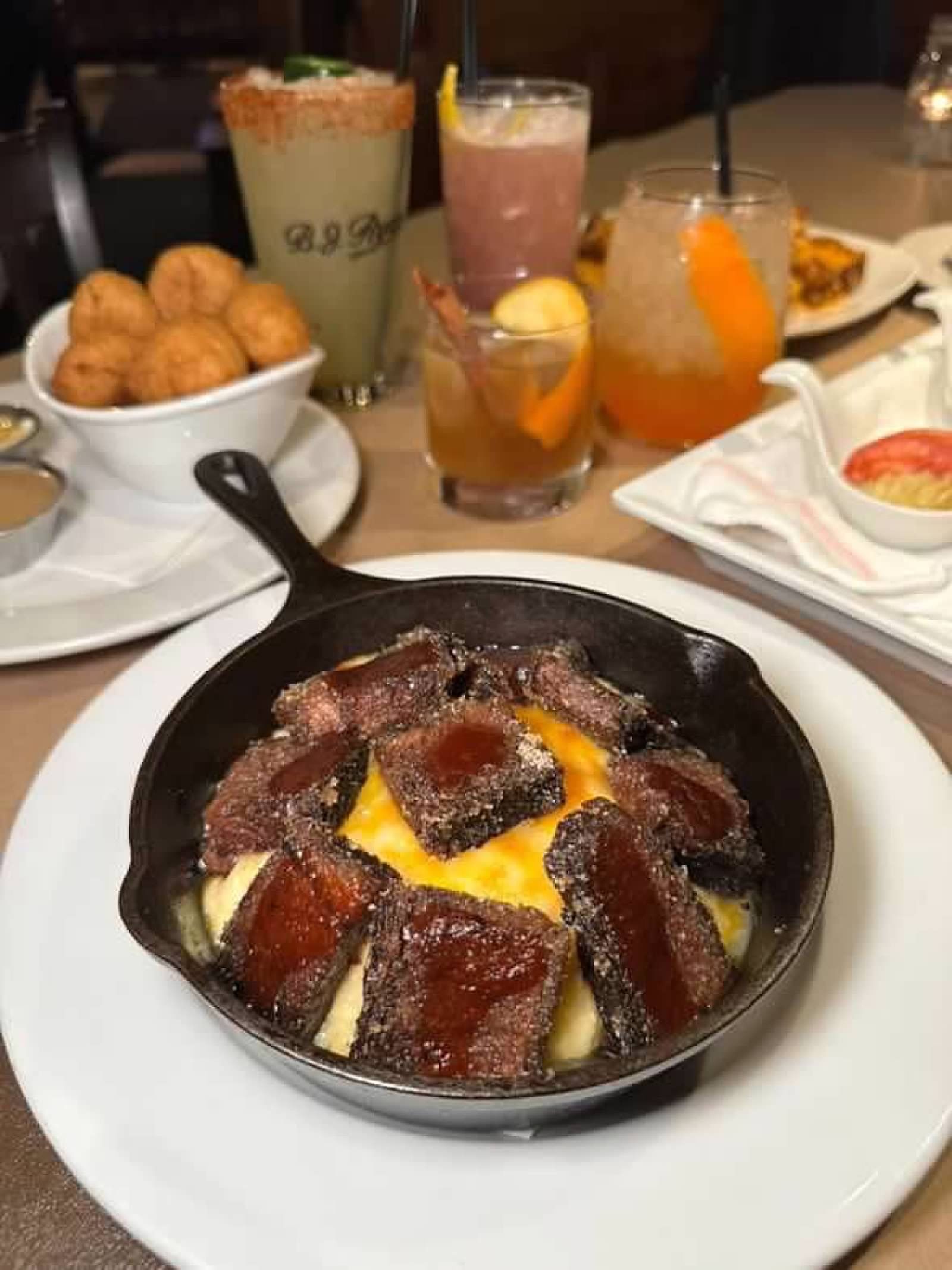 A cast iron skillet with sliced steak over melted cheese, accompanied by a bowl of hush puppies and several cocktails.