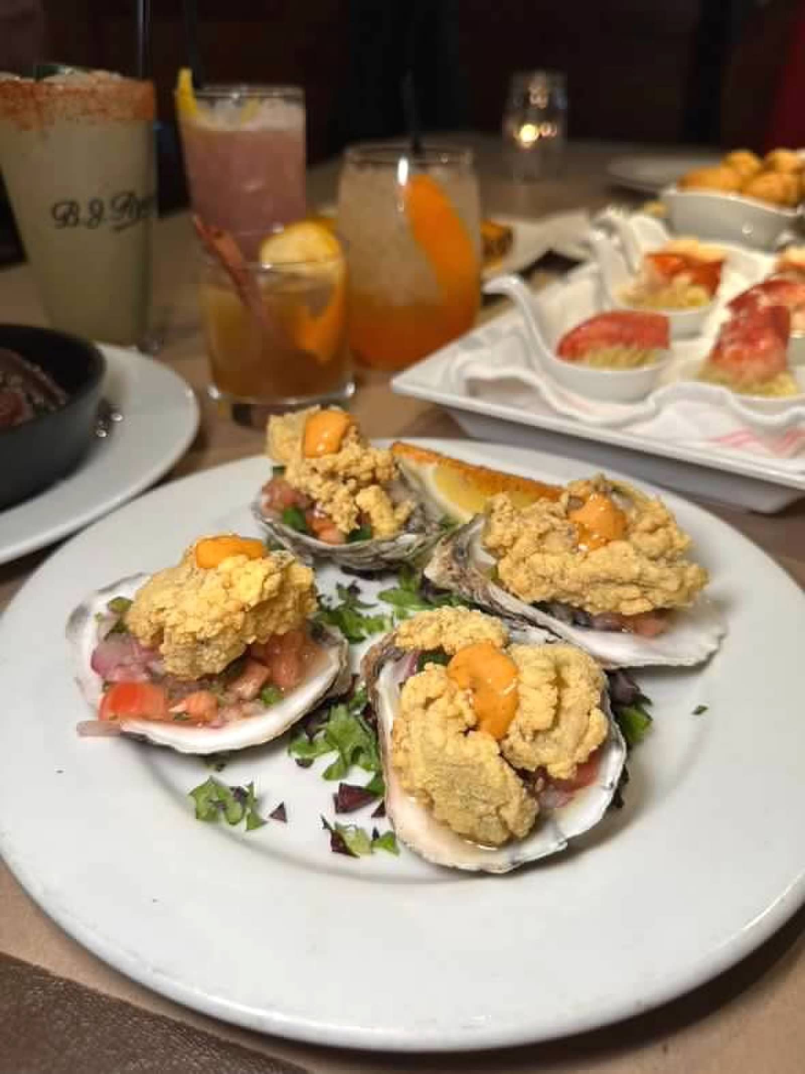 A plate of oysters topped with a creamy garnish and salsa, served on a restaurant table with several colorful cocktails.