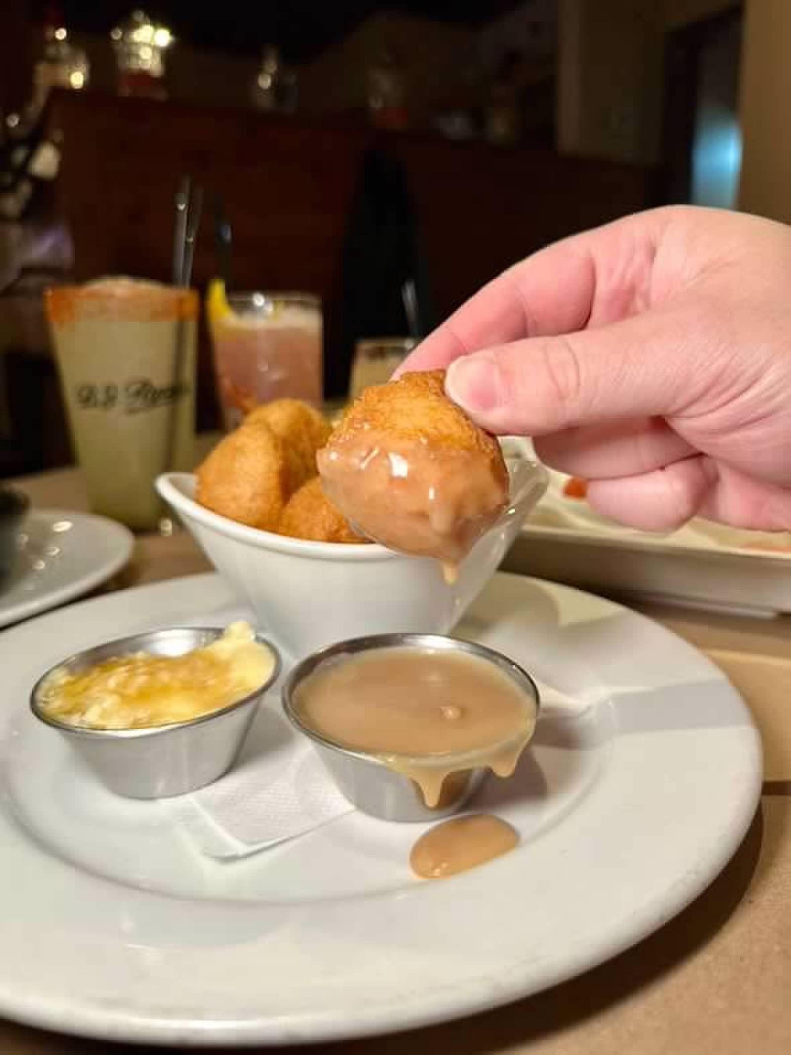 A hand holds a golden-brown appetizer over a white bowl on a plate with two small metal cups of dipping sauce.