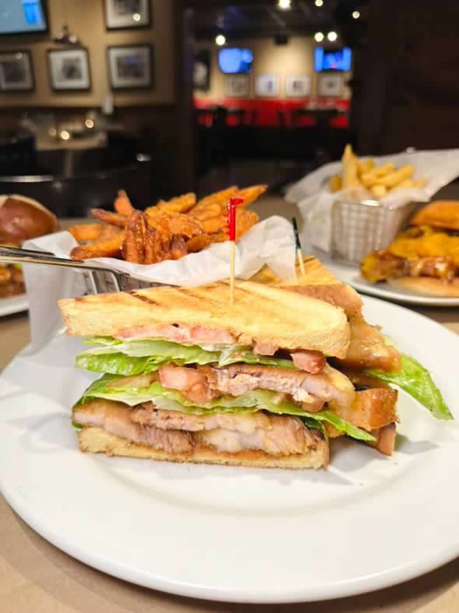 A double-decker grilled chicken sandwich with lettuce and tomato on a plate with sweet potato fries in a restaurant.