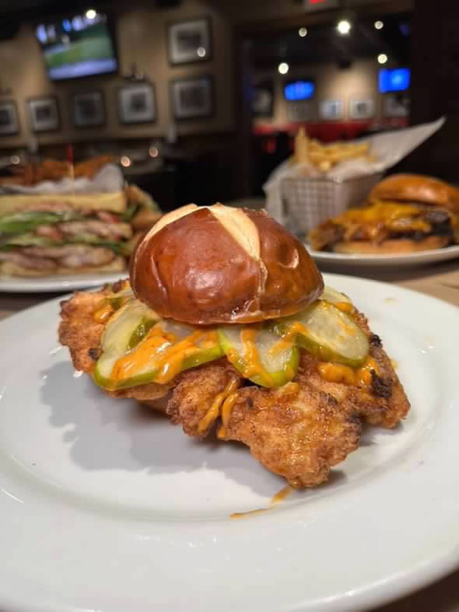 A fried chicken sandwich on a pretzel bun with pickles and sauce, served on a white plate in a restaurant setting.