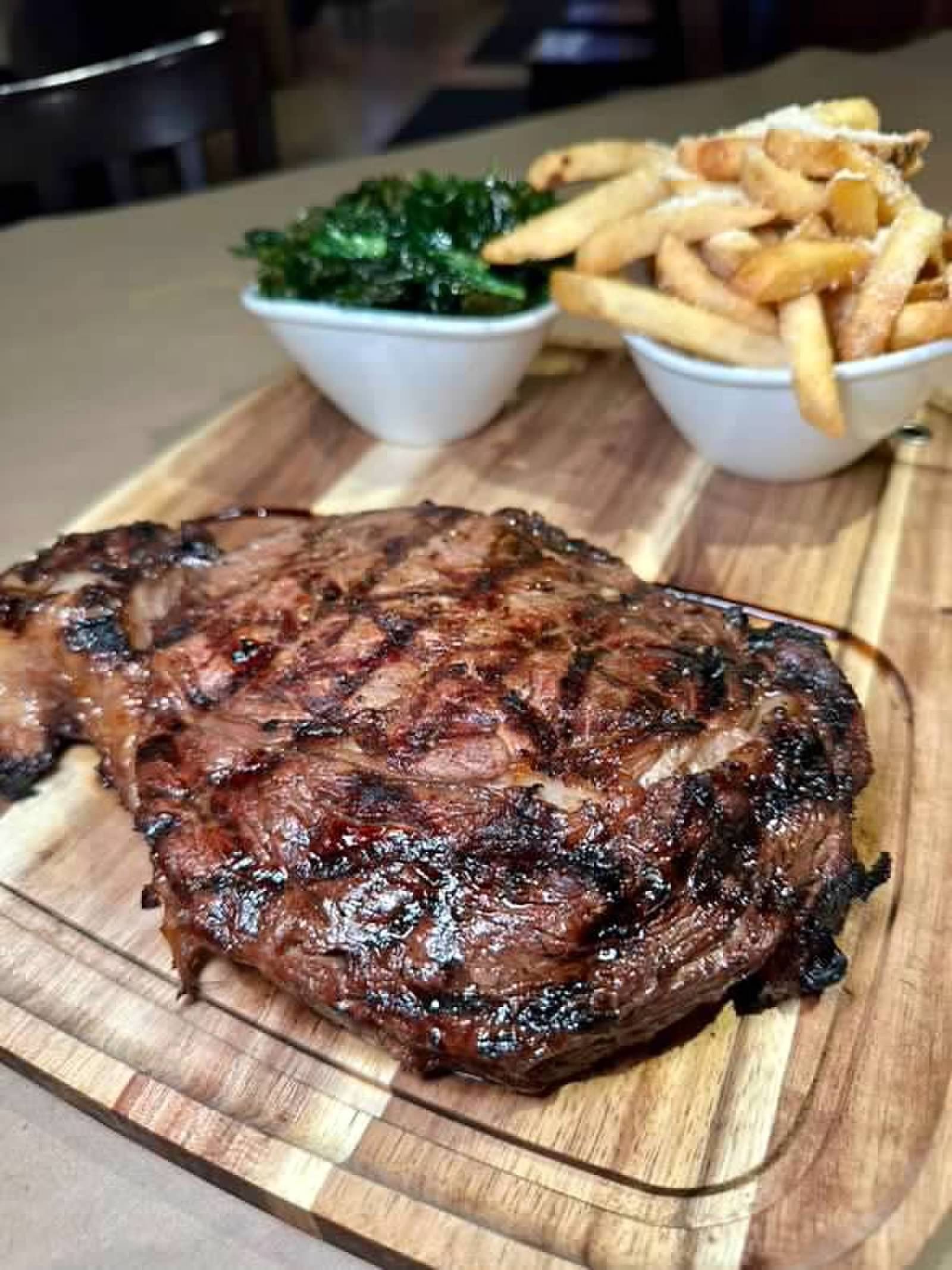 A grilled ribeye steak on a wooden board with small bowls of green vegetables and french fries.