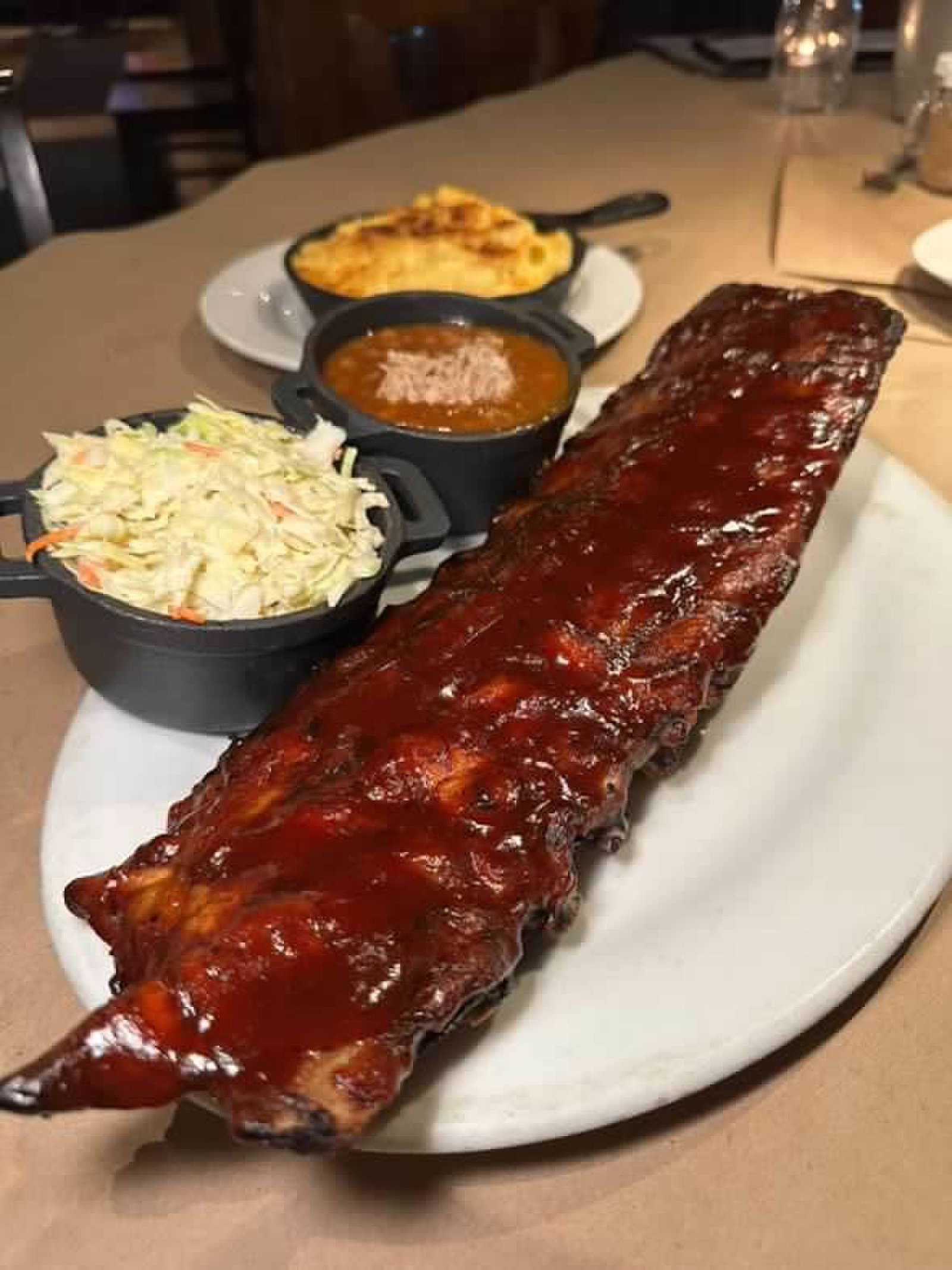 A large rack of glazed barbecue ribs served on a white platter with sides of coleslaw, baked beans, and mac and cheese.