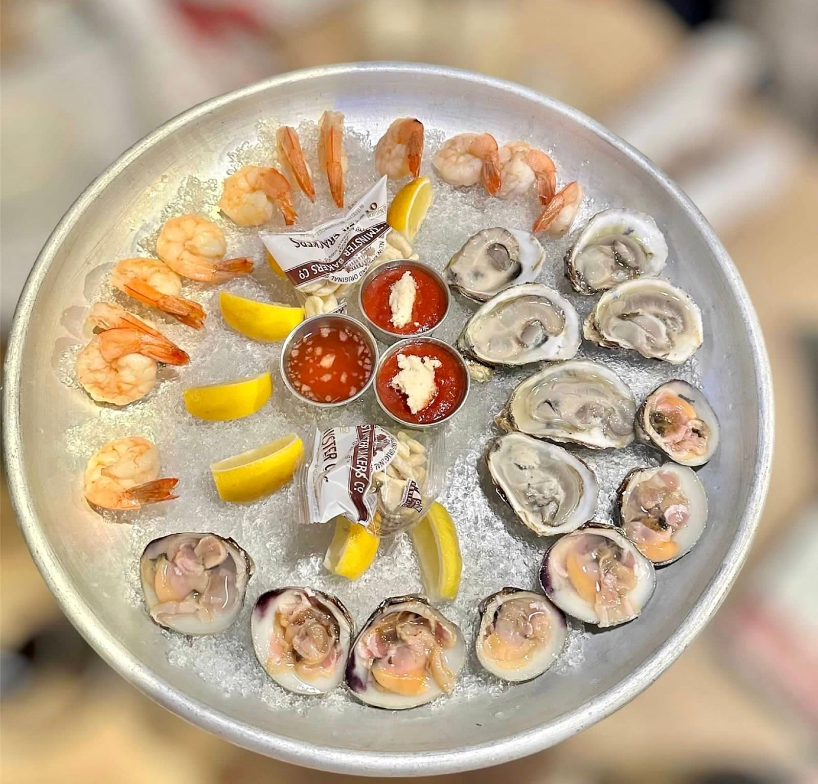 A seafood platter with shrimp, oysters, and clams served on ice with lemon wedges and dipping sauces in a metal tray.