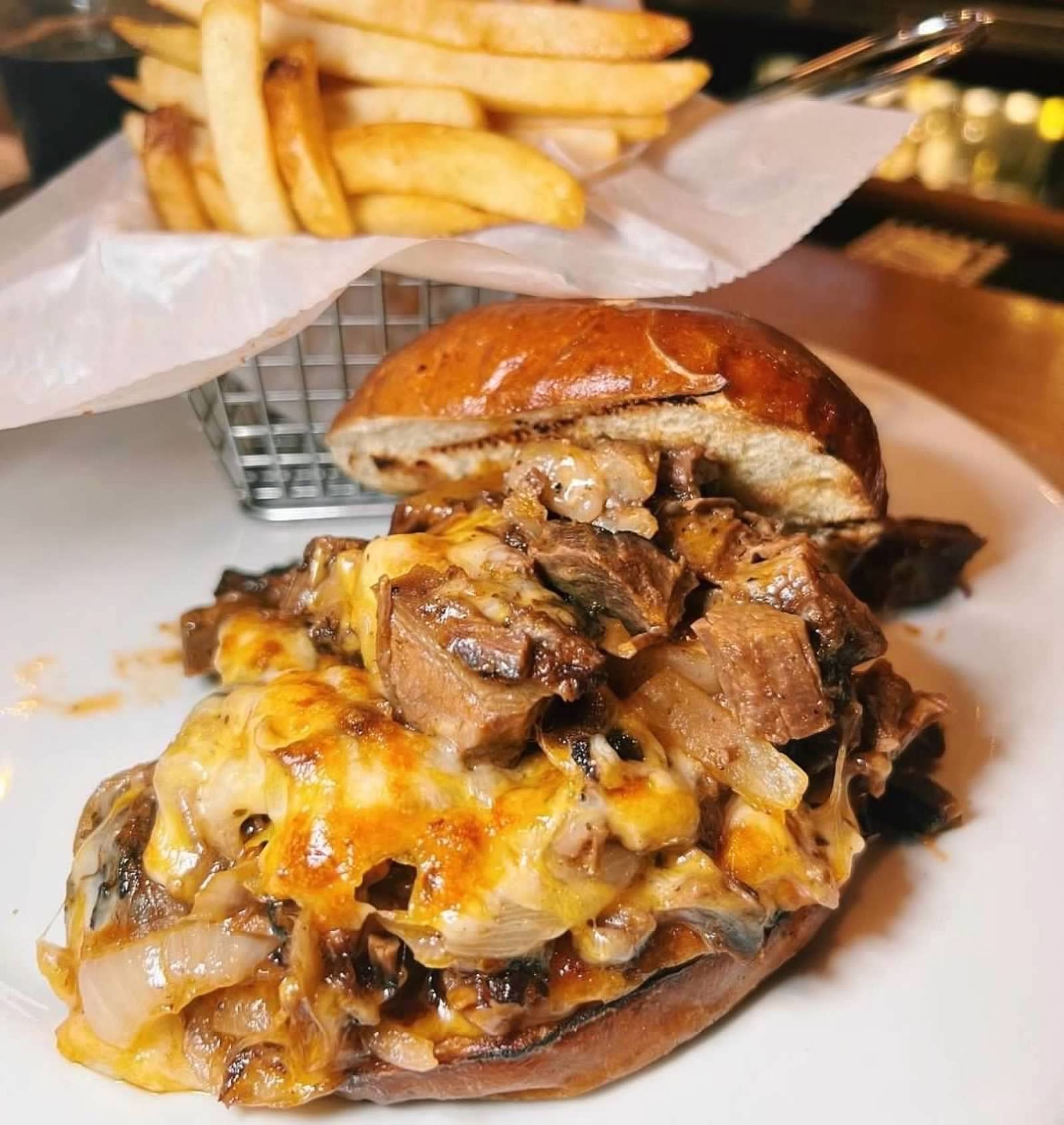 A gourmet roast beef sandwich topped with melted cheese, onions, and mushrooms, served with a basket of golden fries.