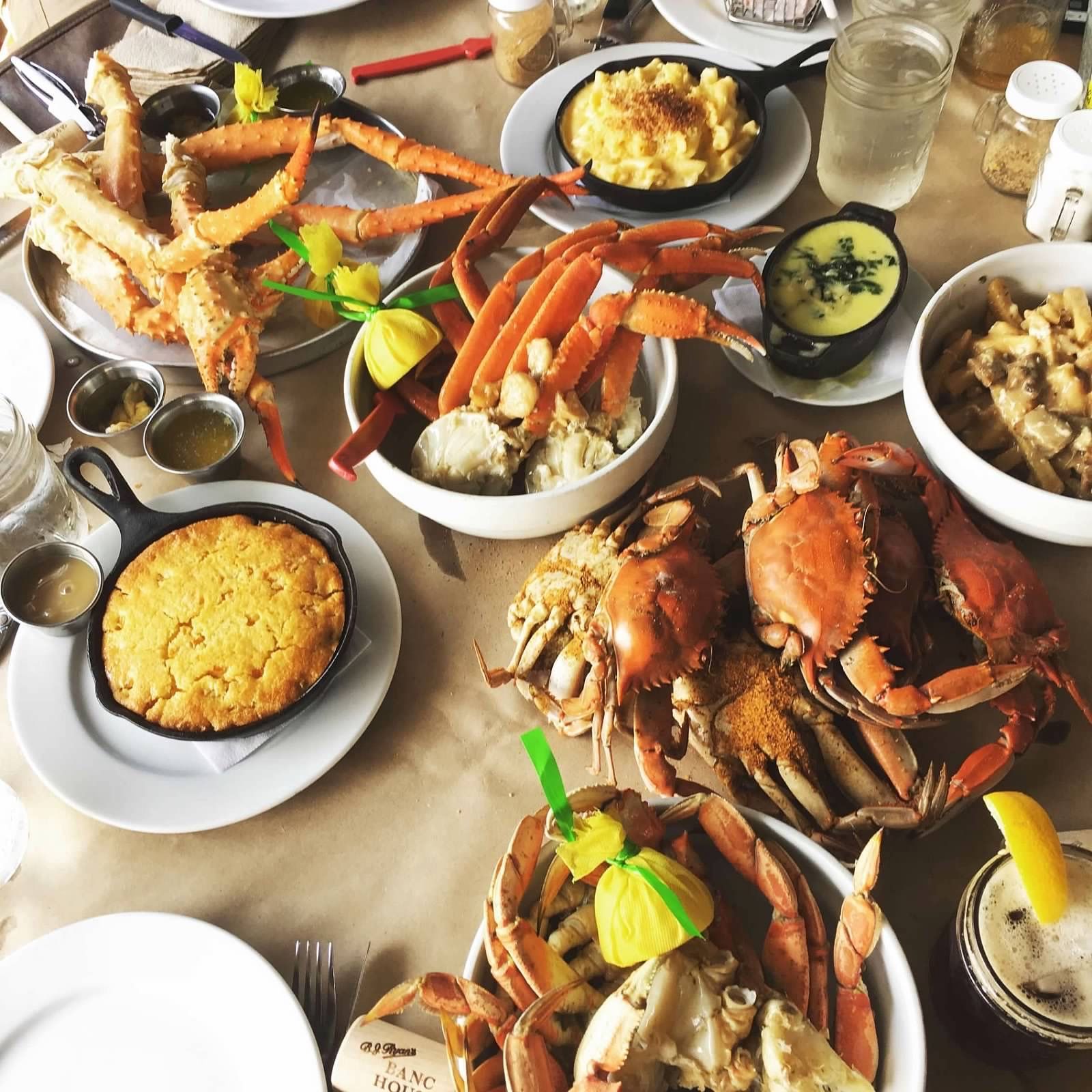 A table spread featuring crab legs, whole crabs, cornbread, mac and cheese, dipping sauces, and drinks on paper.