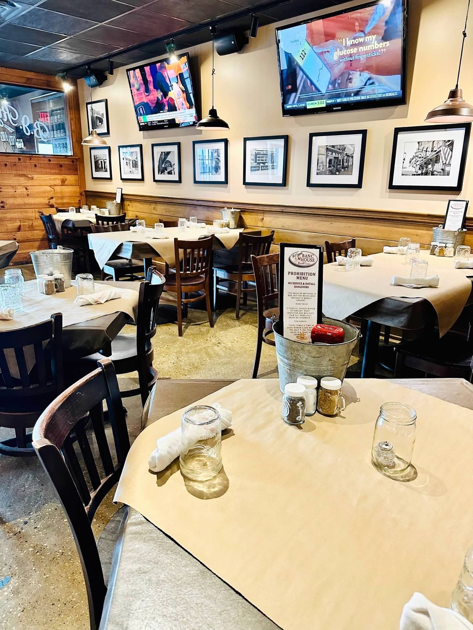 Tables with butcher paper, glasses, and a metal bucket in a restaurant with framed wall art and TVs mounted overhead.