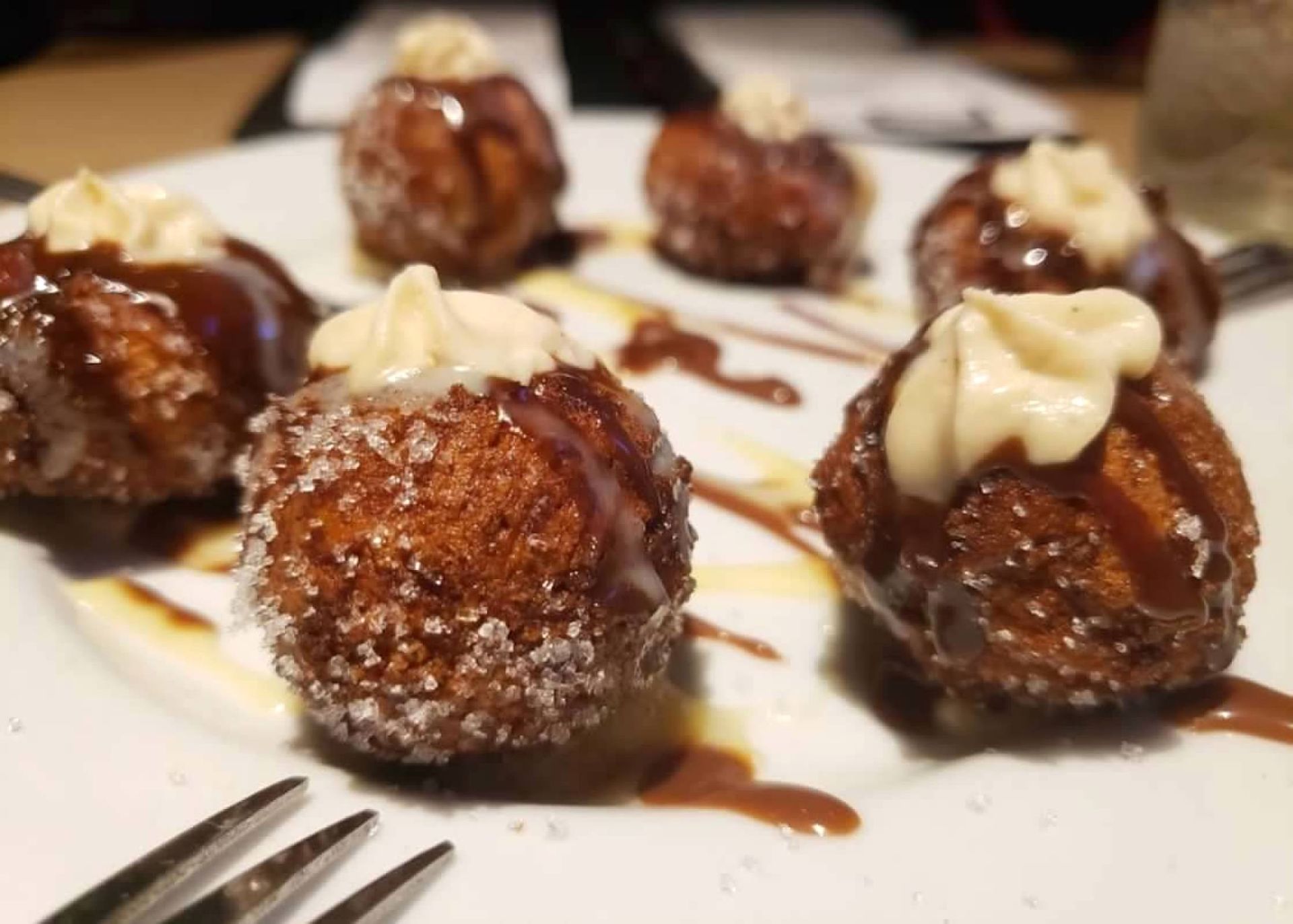 A plate of six fried dough balls sprinkled with sugar, drizzled with chocolate sauce, and topped with cream dollops.