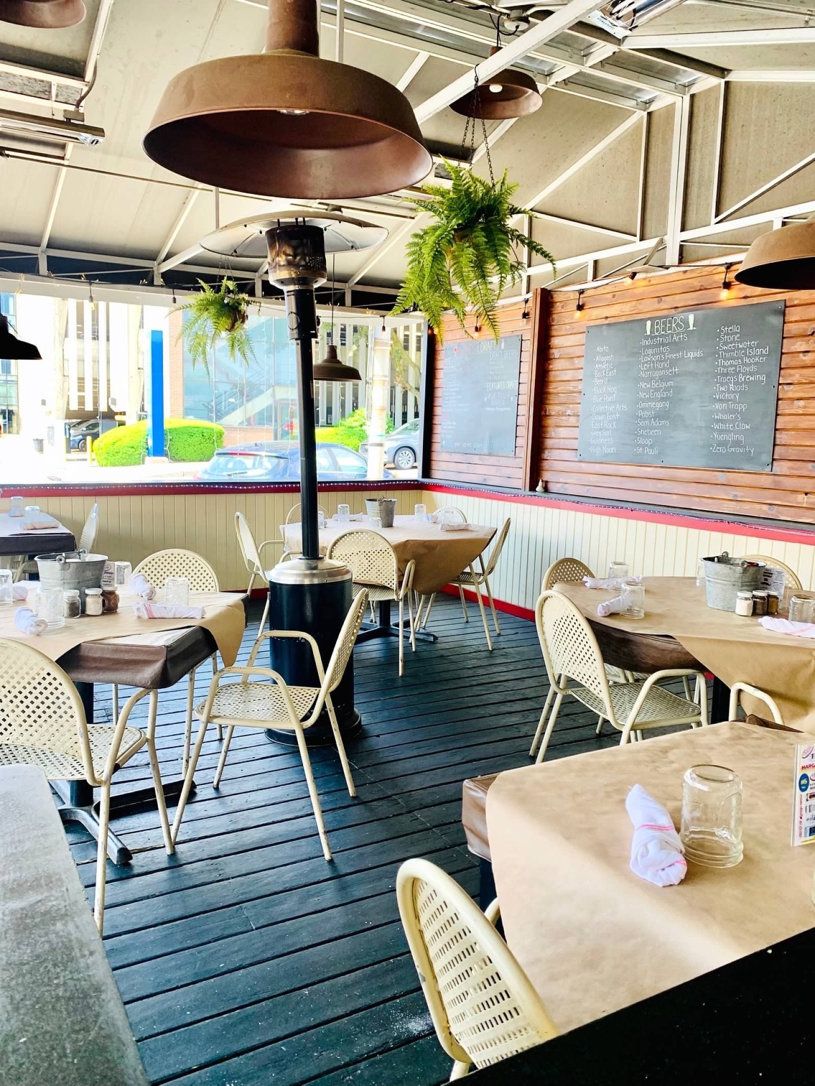 A bright outdoor restaurant patio featuring circular tables, wicker chairs, hanging plants, and an industrial heater.