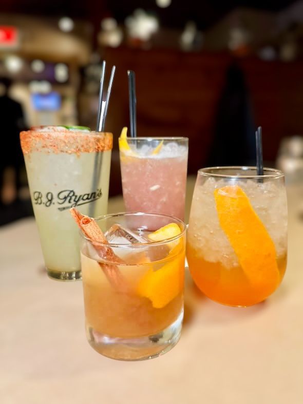 Four assorted cocktails on a bar top, including a tall glass with a salted rim and three shorter glasses with garnishes.