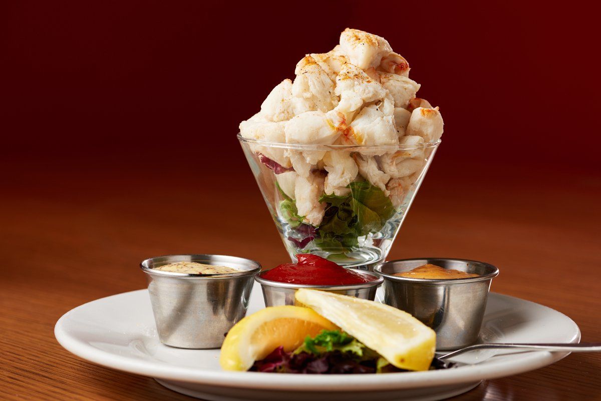 A crab cocktail in a glass on a plate with three metal dipping cups, lemon wedges, and greens against a maroon background.