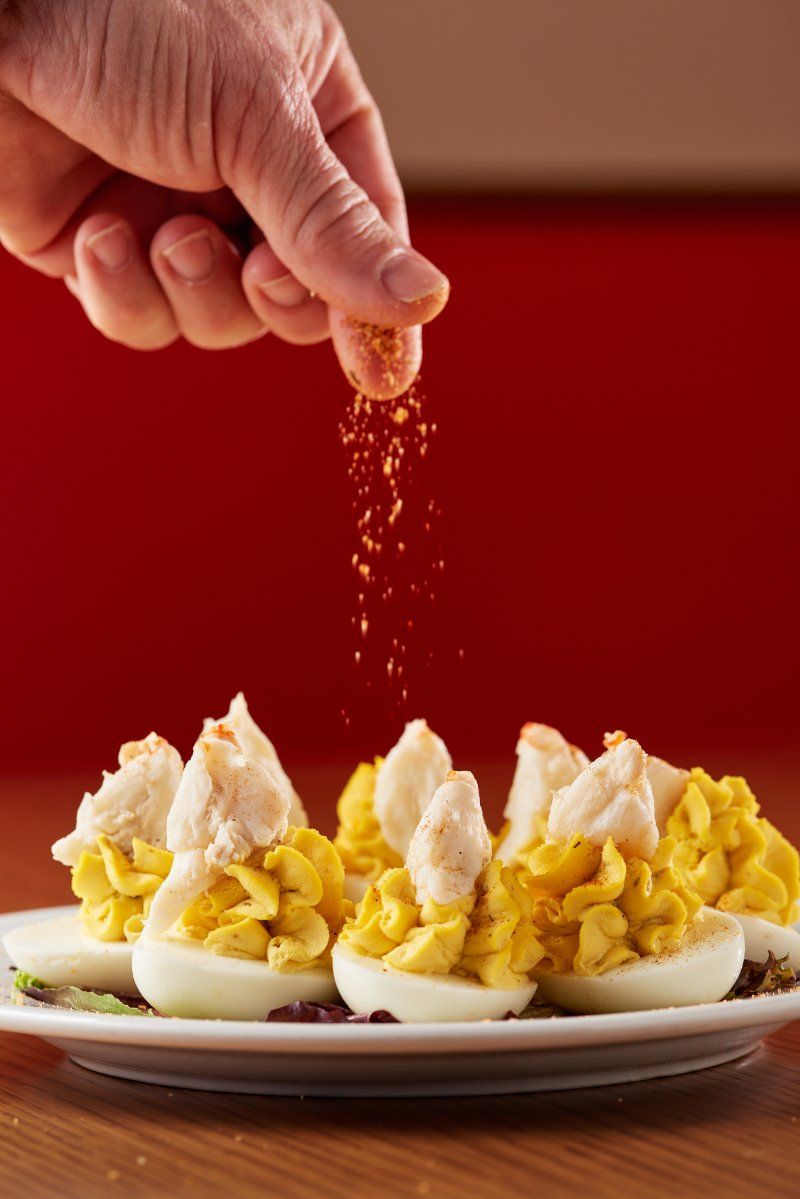 A hand sprinkles paprika over a plate of deviled eggs topped with crab meat, set against a solid red background.