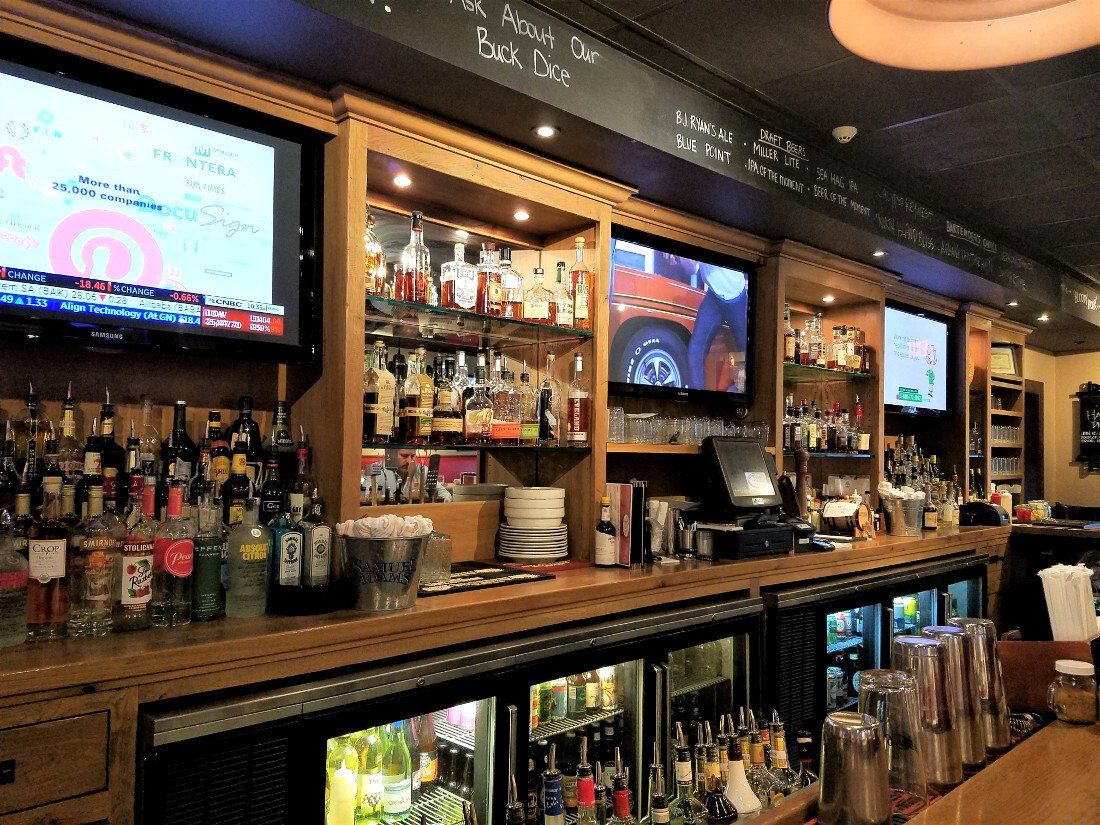 Well-lit bar with shelves of liquor bottles, televisions, and under-counter refrigerators.