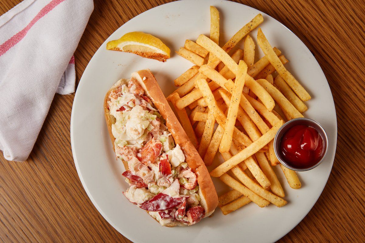 A lobster roll served on a white plate with a side of french fries, a lemon wedge, and a small container of ketchup.