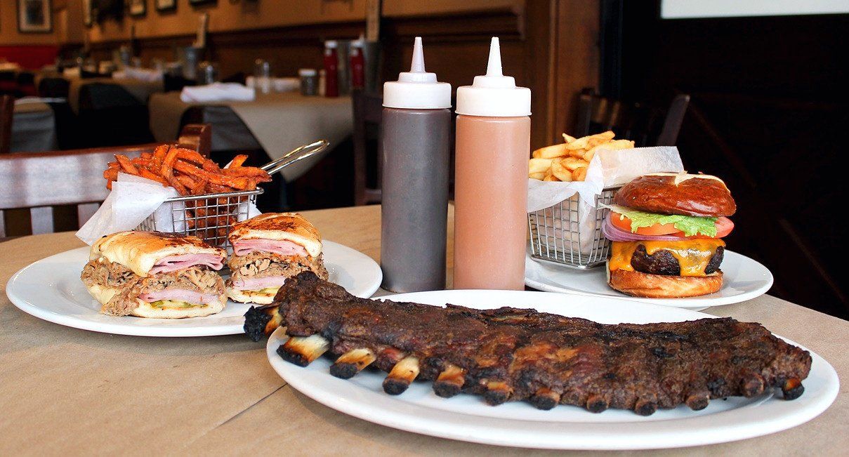 A table with ribs, a burger with fries, and a sandwich with sweet potato fries, with two squeeze bottles in the center.