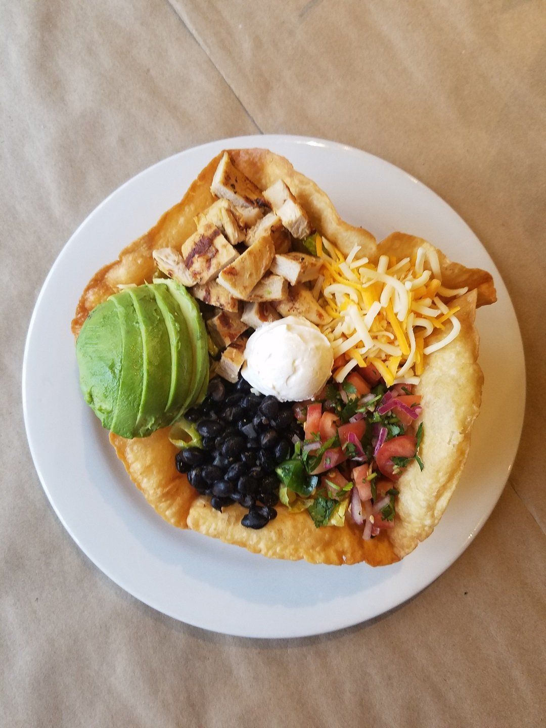 A taco salad in a crispy shell, topped with grilled chicken, sliced avocado, black beans, salsa, cheese, and sour cream.