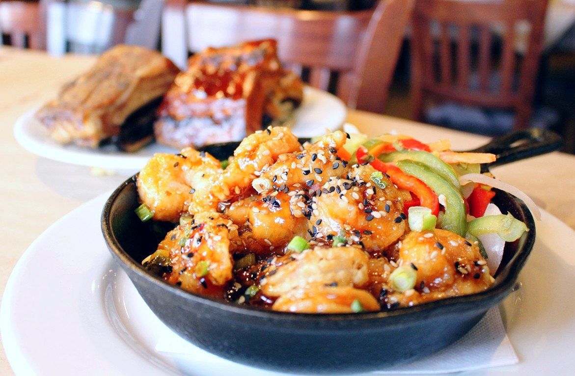 A black cast-iron skillet filled with glazed, sesame-topped shrimp and sliced vegetables, set on a restaurant table.