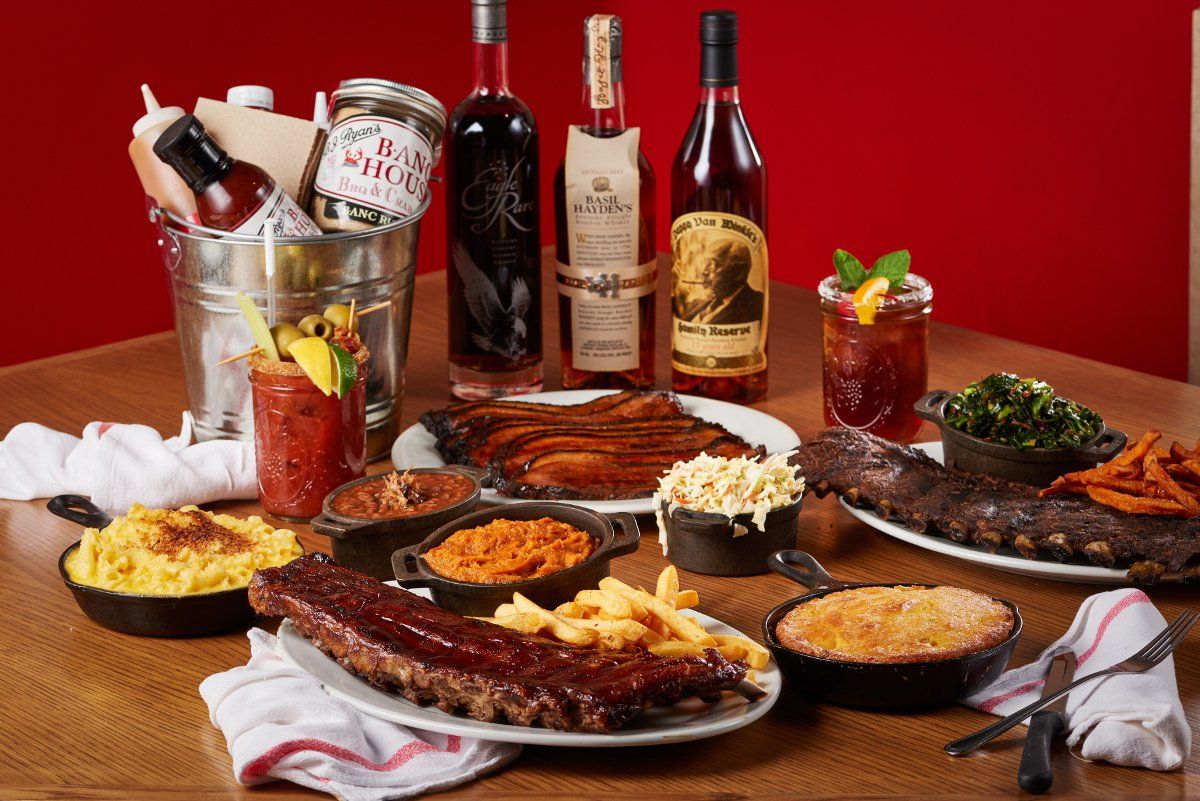 A table spread features BBQ ribs, mac and cheese, various sides, sauces, and cocktail bottles against a red background.