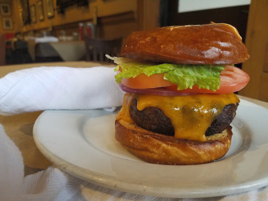 A cheeseburger with lettuce, tomato, and red onion on a pretzel bun, served on a white plate in a restaurant.