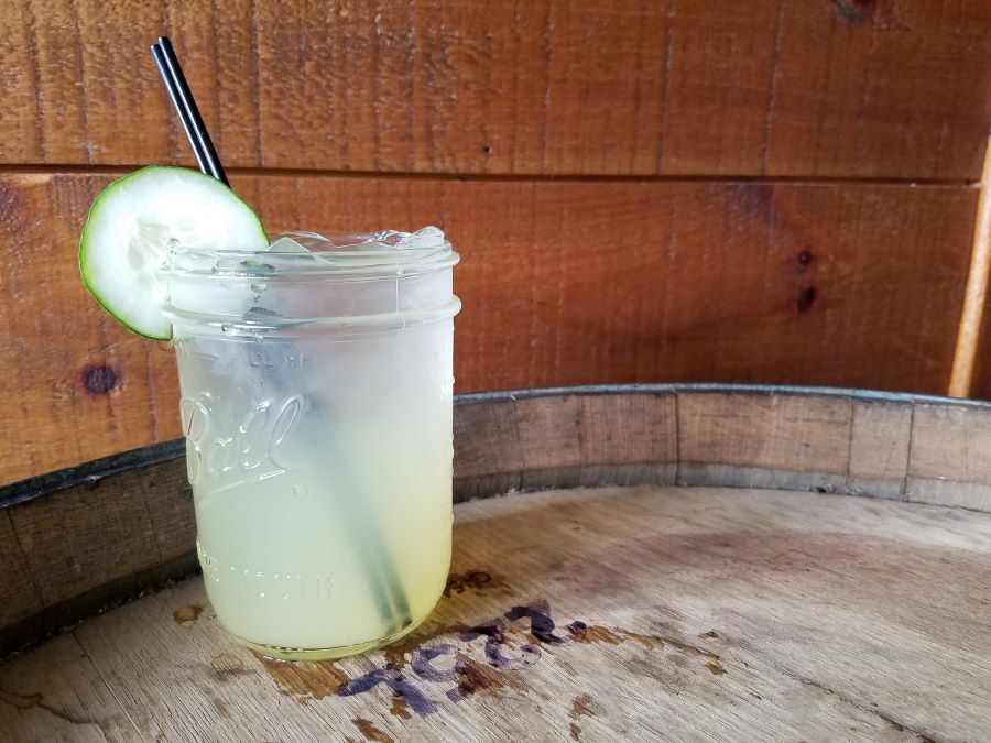 A glass mason jar filled with a pale yellow cocktail, garnished with a cucumber slice, sitting on a wooden barrel.