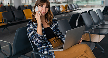 Woman using a laptop and phone in an airport waiting area, seated among rows of chairs.