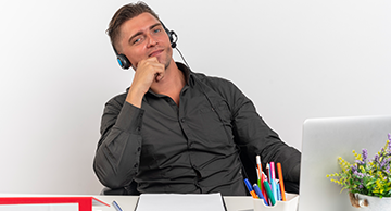 Man wearing a headset sits at a desk, looking thoughtful beside a laptop and pencil holder.