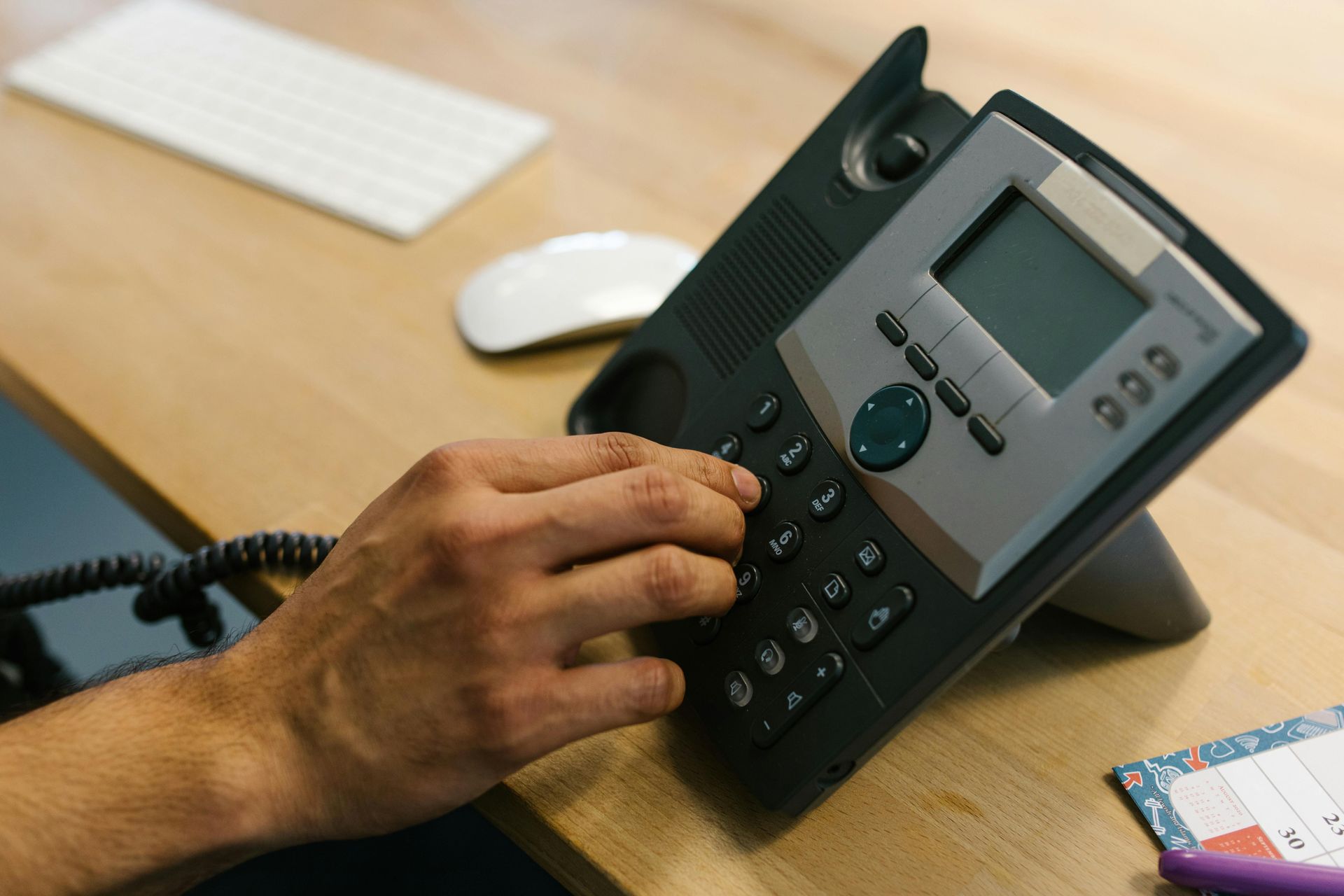 Hand dialing a black office desk phone on a wooden desk with a keyboard nearby