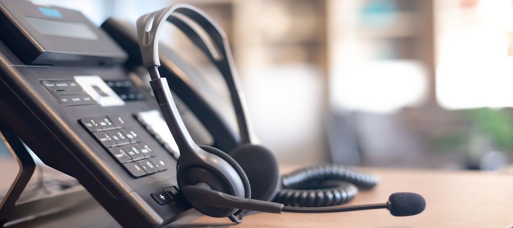 Office desk phone with headset on a wooden table, in a bright blurred workspace