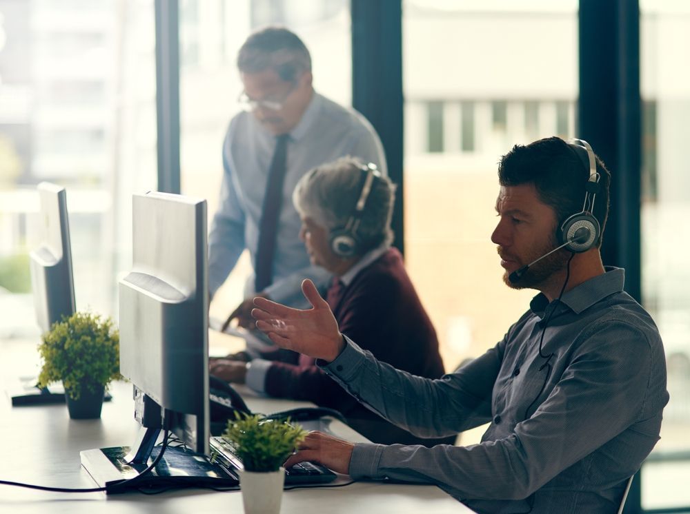 Office workers collaborating at computers, one wearing a headset and gesturing while speaking.