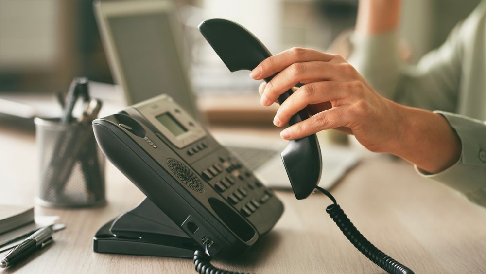 Hand holding a black office phone handset above a desk phone in a bright office