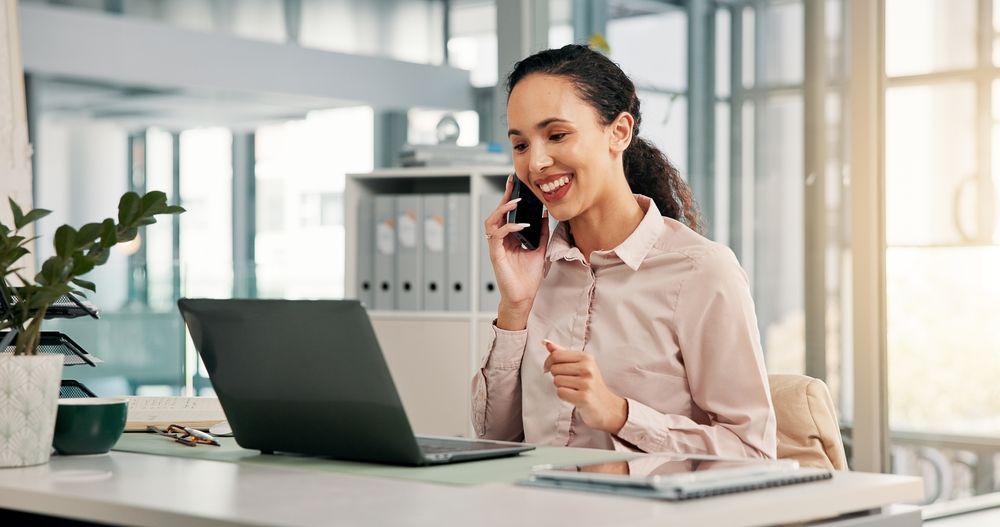 Woman smiling on a phone call at a desk with a laptop in a bright office