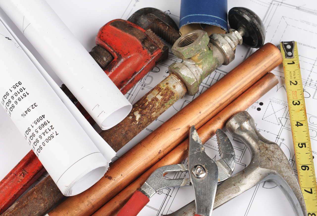Plumbing tools and blueprints scattered: wrench, hammer, pipe, measuring tape, and rolled plans.