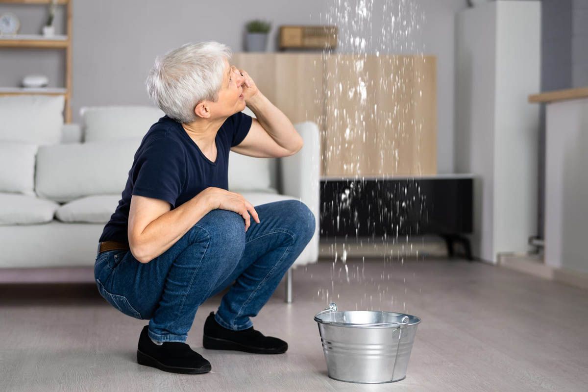 Woman squatted under a ceiling leak, using hand to call Pat's Plumbing, with bucket on floor.
