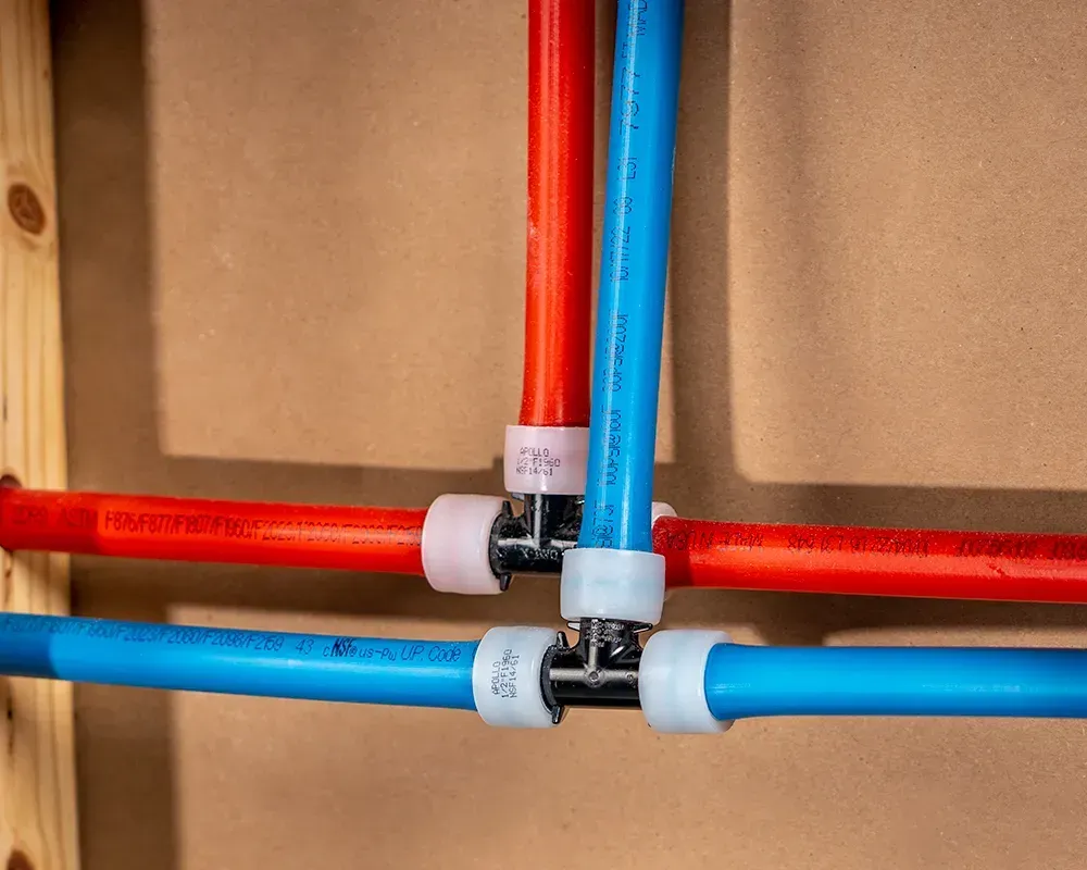 Pipes with T-fittings, red and blue, for hot and cold water plumbing against a beige wall.