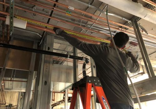 Two people lift and maneuver a cylindrical water heater in a gray-walled room.