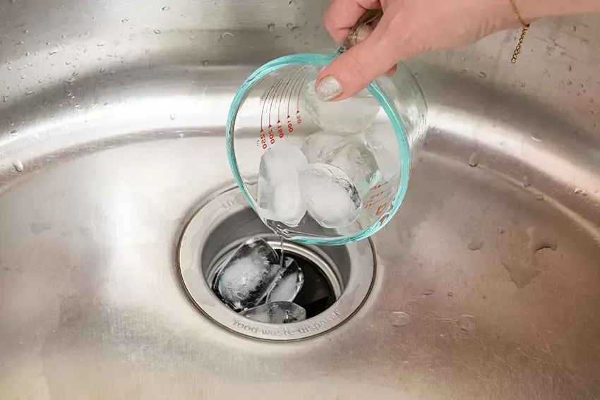 Hand pouring ice cubes from a measuring cup into a kitchen sink disposal.