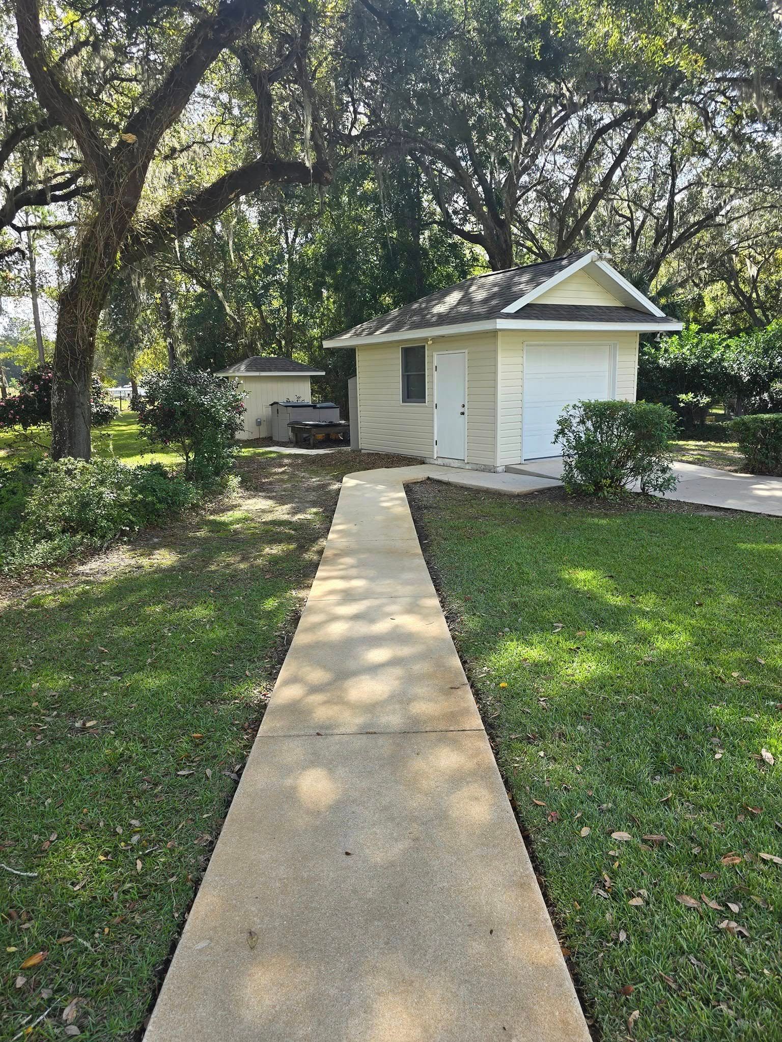 A concrete walkway leading to a small white house surrounded by trees.