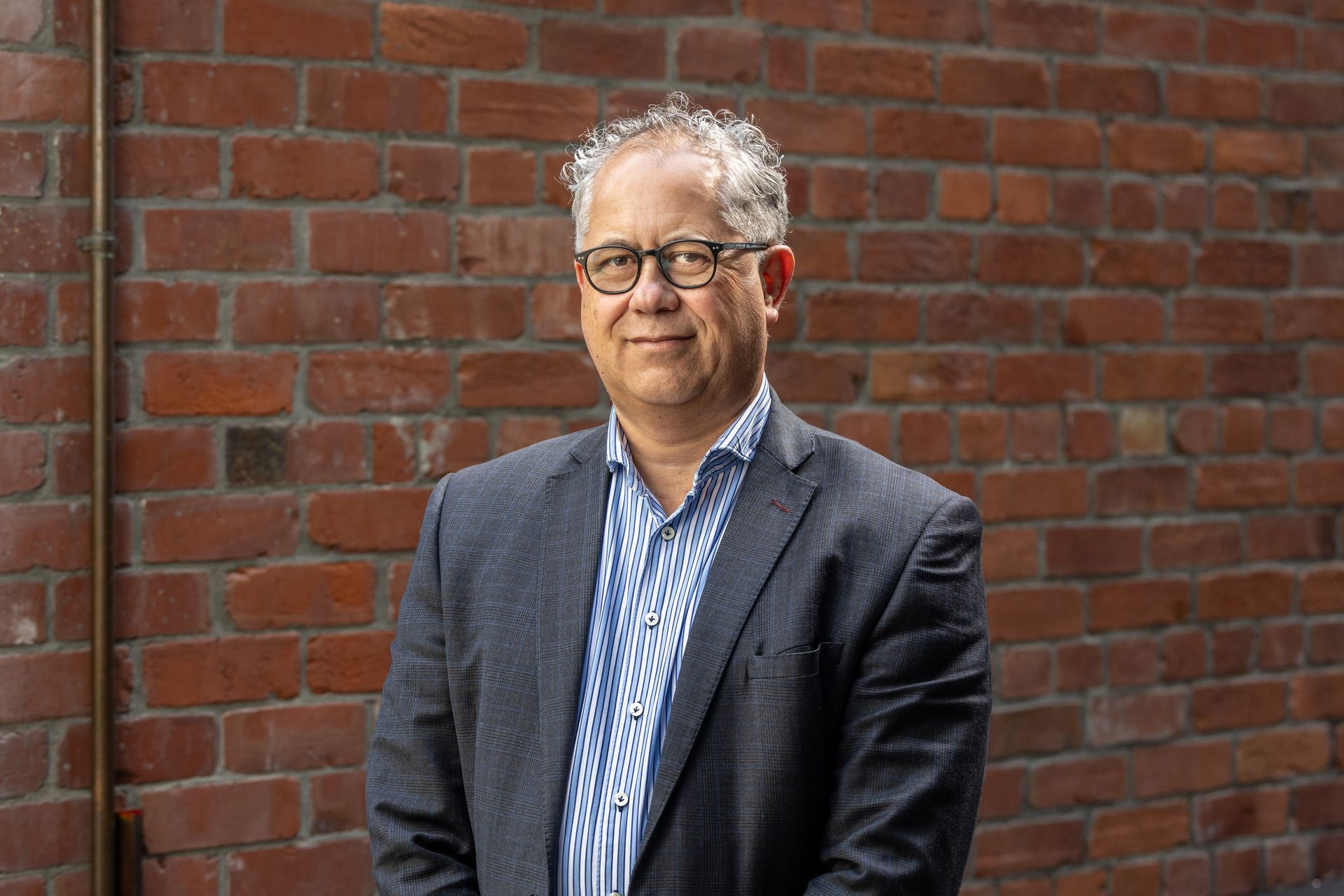 A man in a suit and glasses is standing in front of a brick wall.