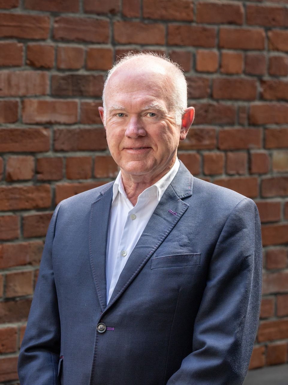 A man in a suit and white shirt is standing in front of a brick wall.