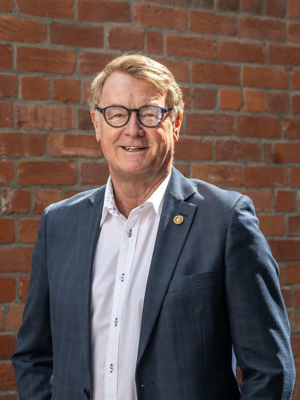 A man in a suit and glasses is standing in front of a brick wall.