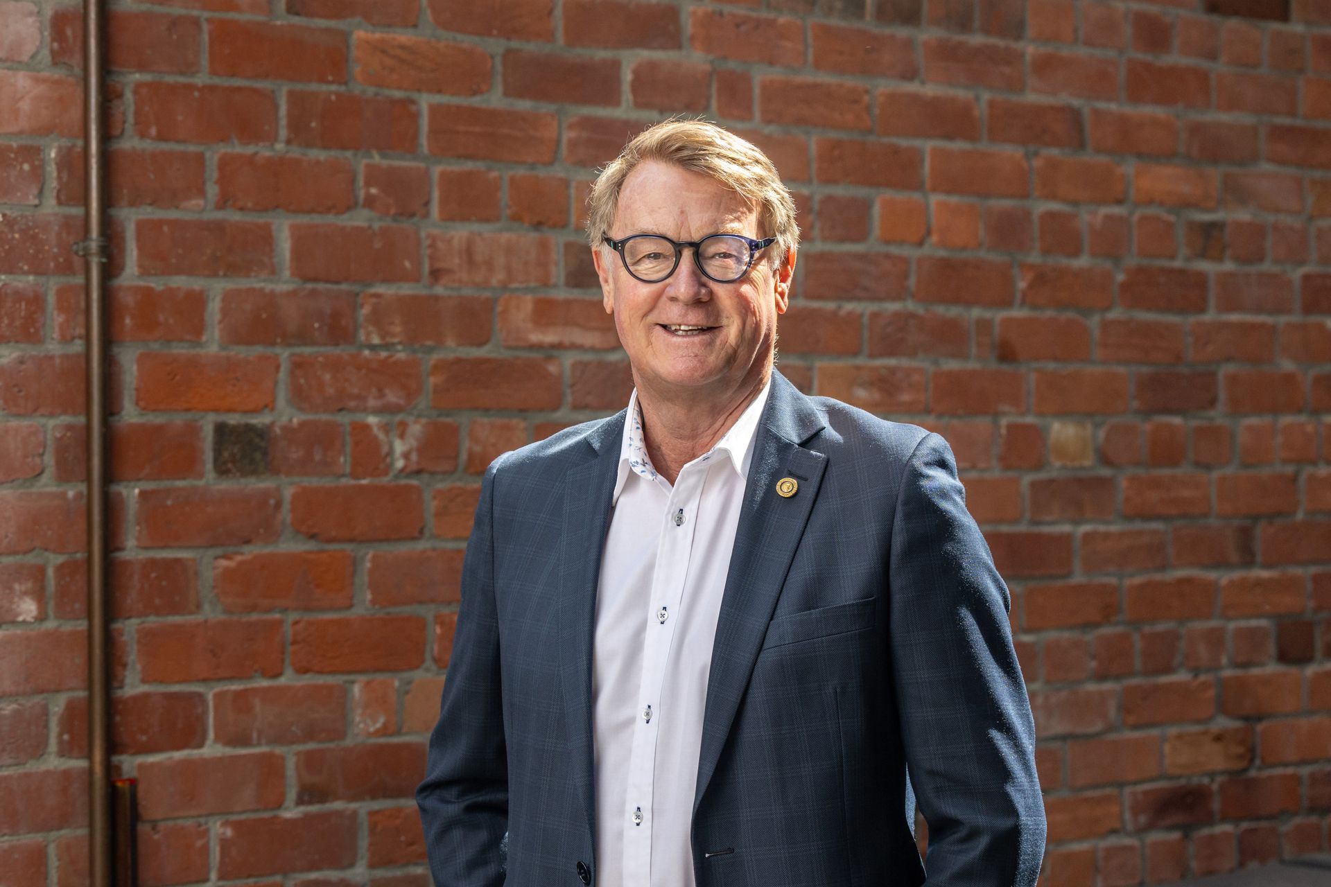 A man in a suit and glasses is standing in front of a brick wall.