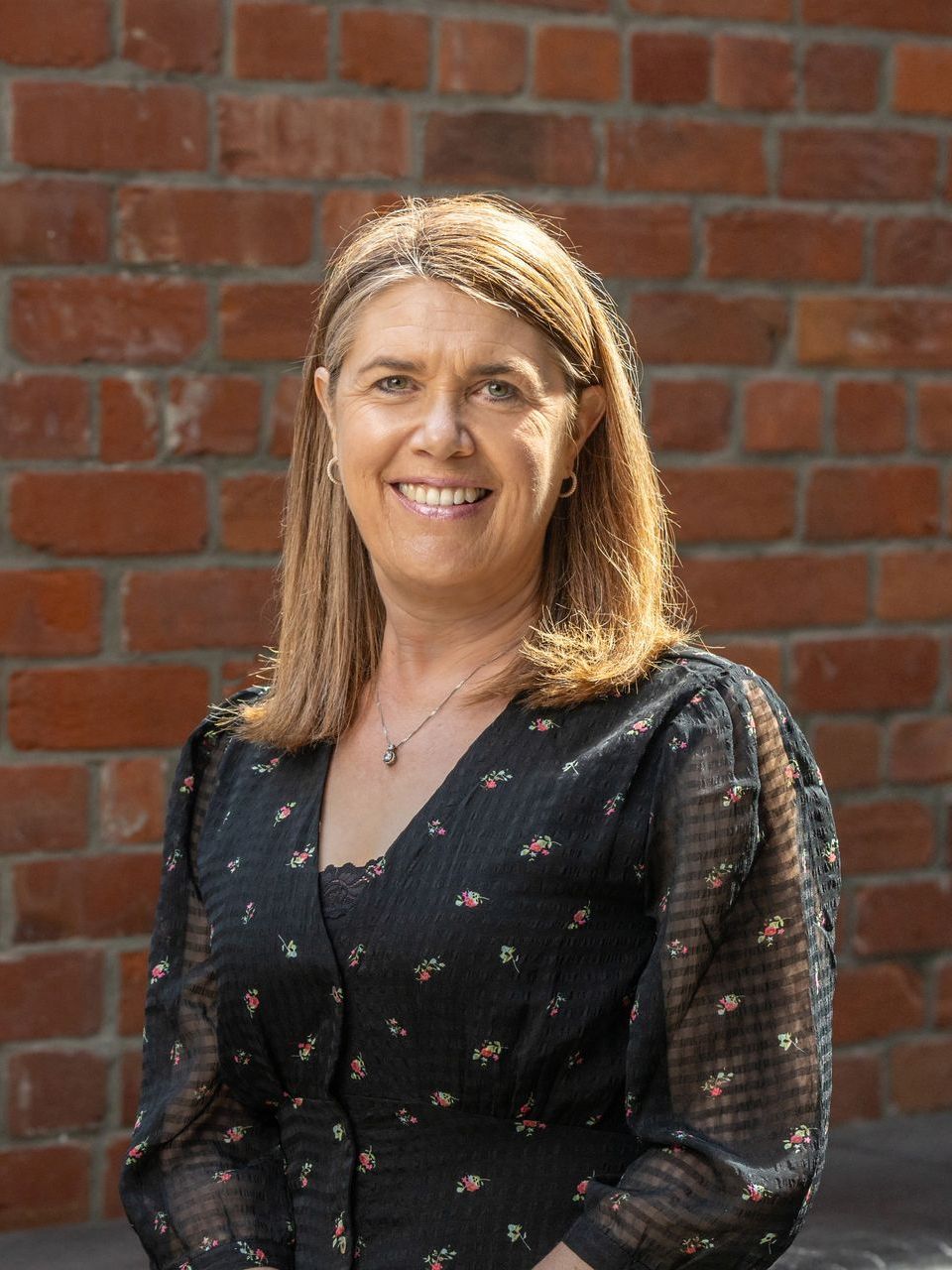 A woman in a black dress is smiling in front of a brick wall.