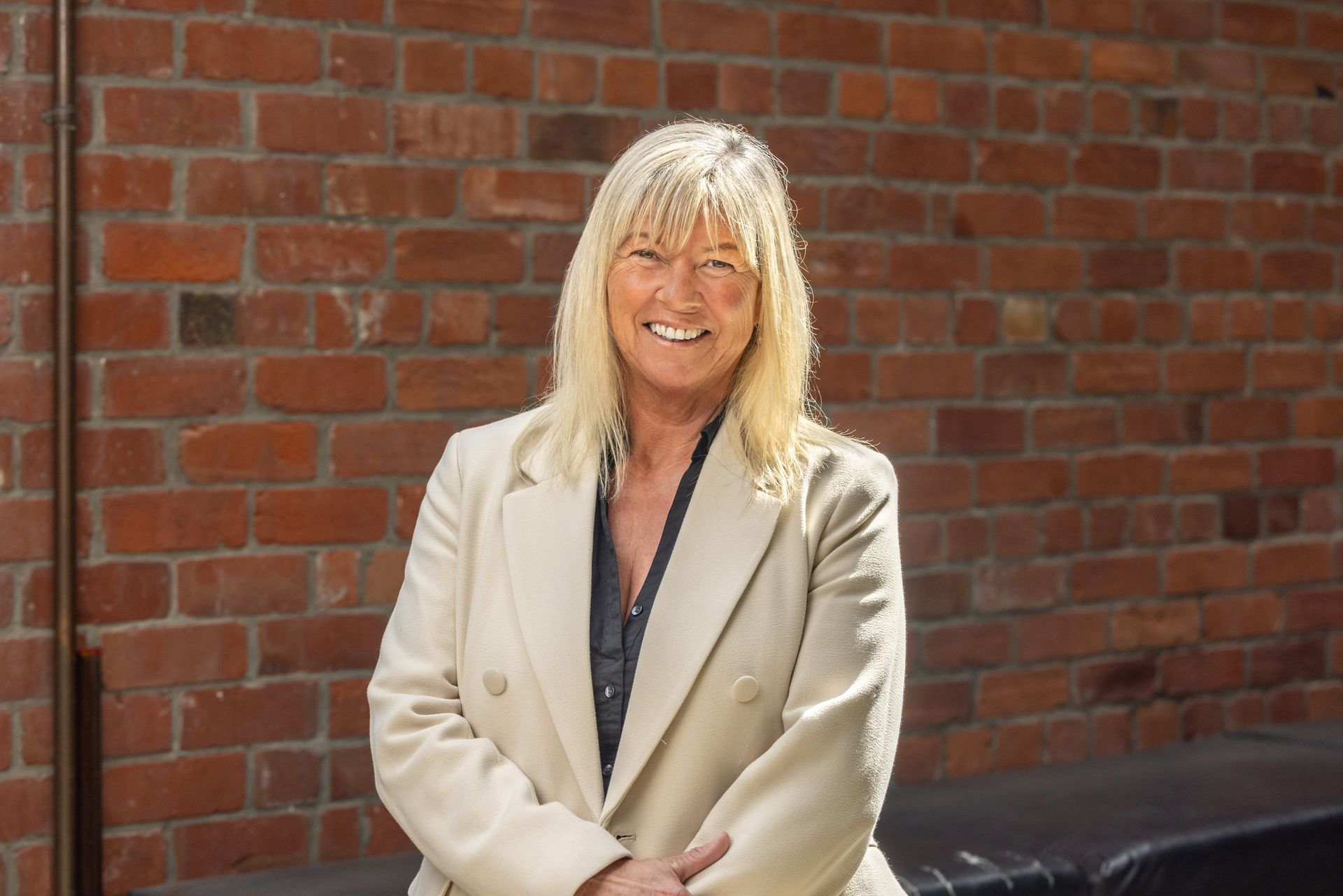 A woman in a white jacket is standing in front of a brick wall.