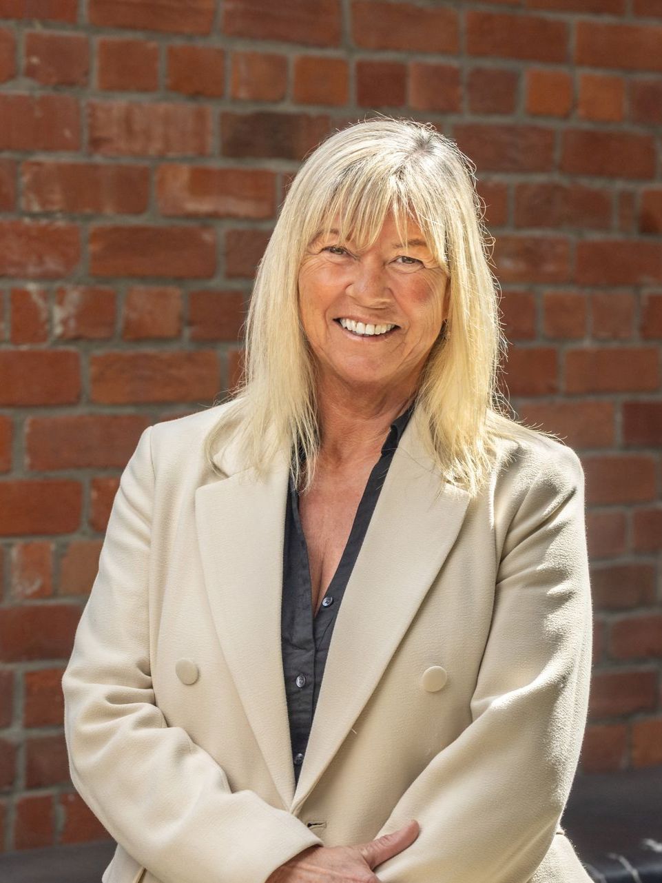 A woman in a white coat is smiling in front of a brick wall.