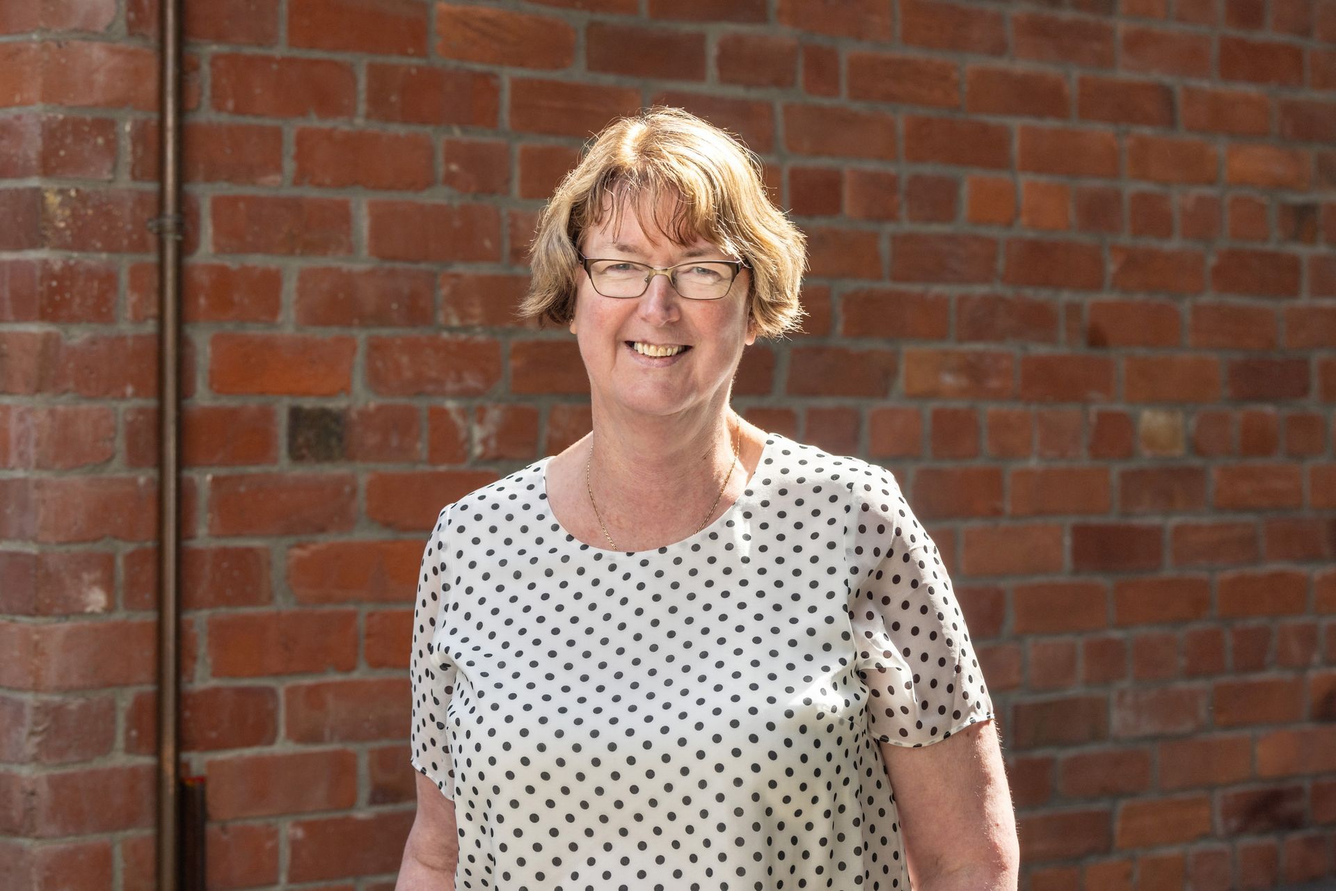 A woman wearing glasses and a polka dot shirt is standing in front of a brick wall.