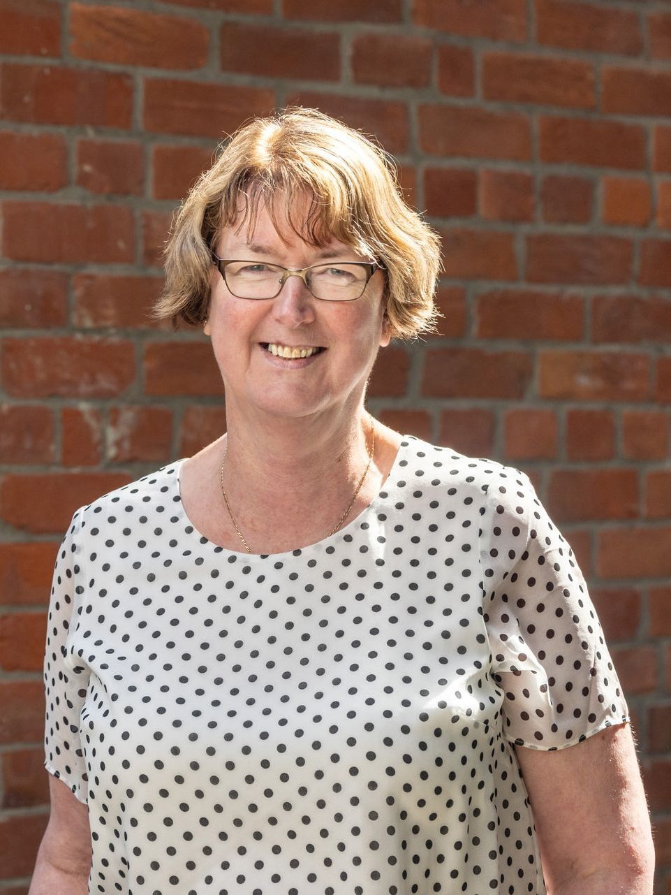A woman wearing glasses and a polka dot shirt is smiling in front of a brick wall.