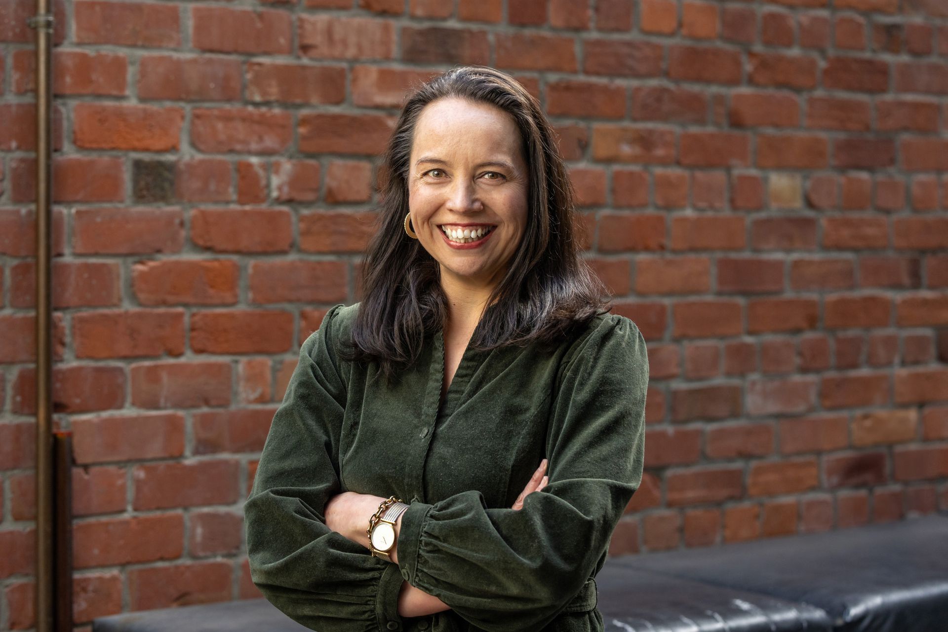 A woman in a green jacket is standing in front of a brick wall with her arms crossed.