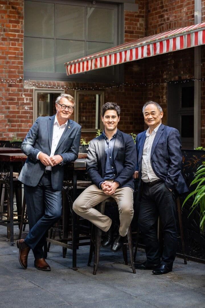 Three men are standing next to each other in front of a brick building.